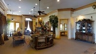 This single-story space showcases a richly appointed interior with a rustic yet refined aesthetic. The main living area draws the eye with vaulted ceilings fitted with multiple ceiling fans and recessed lighting, creating a bright and airy atmosphere. Warm, neutral-toned walls are trimmed with natural wood crown molding, adding architectural character throughout. A stone fireplace serves as a focal point in the living area, flanked by draped windows that allow natural light to filter in. The space is furnished with a plush sofa, area rug, and an ornately carved wooden credenza featuring a scenic landscape design, contributing to the room's decorative richness. Lush indoor trees and elaborate floral arrangements add vibrant natural elements to the interior. An open passageway framed with wood trim leads to an adjacent game room, visible through the doorway, where a pool table takes center stage under warm lighting. The seamless flow between spaces, combined with the layered decorative details, gives the overall interior a welcoming and well-curated character suited for both relaxation and entertainment.