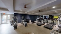This fitness center offers a comprehensive, thoughtfully designed workout facility spread across a single floor. The space is divided into distinct functional zones, each tailored to a different style of exercise. The main cardio and strength training area features speckled terrazzo-style flooring and is outfitted with multiple treadmills, elliptical machines, and a full dumbbell rack, all set against a bold navy blue accent wall that adds a sleek, modern contrast to the light-colored ceiling with recessed lighting. Full-length mirrors line one wall, enhancing the sense of openness and allowing users to monitor their form. A separate studio room provides a dedicated space for group fitness, yoga, or stretching, finished with warm wood-look plank flooring and a vibrant yellow-green accent wall. This studio includes built-in wooden cabinetry stocked with medicine balls, resistance tools, and other fitness accessories, along with a wall-mounted flat-screen TV and expansive floor-to-ceiling mirrors. Glass doors connect the two areas, maintaining visual openness while allowing acoustic separation. The facility is accessible via double entry doors and benefits from natural light near the entrance, creating a welcoming and energizing atmosphere throughout.