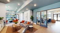 Located in Huntsville, Alabama, this thoughtfully designed single-floor amenity space offers a generous, open layout that seamlessly blends social, recreational, and professional environments. The interior is defined by a cohesive color palette of steel blue accent walls, crisp white trim, and warm light wood flooring that flows throughout the entire space.

The main lounge area features plush seating with vibrant orange accent pillows, anchored by a woven area rug and complemented by natural greenery. Adjacent to it, a striking textured geometric feature wall serves as the backdrop for a mounted flat-screen TV, a sleek wet bar with white cabinetry, and a built-in beverage refrigerator—ideal for entertaining. Large windows in this zone invite natural light and offer pleasant outdoor views.

A dedicated game room includes a full-size billiards table and casual dining seating, decorated with sculptural wall art and tall potted trees that add a refreshing, organic touch. A separate dining nook with a dark wood table and framed gallery wall provides a quieter setting for meetings or collaborative work.

Glass-fronted private office pods offer semi-enclosed workspaces outfitted with ergonomic seating, providing focused work areas within the broader open-plan environment. A media lounge with swivel club chairs arranged around a wall-mounted TV rounds out the amenities. Recessed lighting throughout ensures a well-lit, welcoming ambiance across every zone.