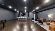 Located at 100 Shepherds Lane in Totowa, NJ, this sophisticated amenity lounge is a generously sized, single-floor space designed for socializing and recreation. The interior is defined by a striking dark navy ceiling that contrasts beautifully with cool grey-blue walls and rich, dark hardwood flooring throughout, creating a refined yet inviting atmosphere.

The space is thoughtfully divided into distinct functional zones. A stylish wet bar area anchors one side of the room, featuring sleek grey cabinetry with white countertops, a gold-accented sink fixture, open display shelving, and a wall-mounted flat-screen TV. A long communal bar-height island with modern white chairs provides ample seating, making it ideal for gatherings or casual dining.

Adjacent to the bar area, a dedicated billiards zone showcases a full-size pool table with a bold red felt surface, positioned beneath recessed lighting. A glass partition wall adds a modern architectural element, visually connecting this zone to the broader space.

The lounge area offers a curated seating arrangement with tufted caramel leather sofas, deep navy velvet accent chairs, woven ottomans, and a soft area rug, all illuminated by a warm floor lamp. Recessed ceiling lights provide even, ambient illumination throughout, enhancing the space's upscale, club-like character.