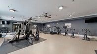 Located at 120 Chubb Ave in Lyndhurst, NJ, this fitness center provides a generously sized, single-floor workout facility designed to accommodate a full spectrum of exercise needs. The space is finished with a neutral-toned resilient floor, crisp white crown molding, and deep charcoal-gray walls that lend a modern, energizing atmosphere throughout.

The cardio zone features multiple treadmills, elliptical trainers, and stationary cycling bikes, all aligned along one wall for an organized and efficient layout. Ceiling fans with flush-mount lighting fixtures ensure consistent airflow and comfort during workouts. Wall-mounted televisions are positioned throughout the space for entertainment during cardio sessions.

The strength training area is equally well-appointed, housing a multi-station cable machine capable of a wide variety of resistance exercises. A dedicated free weights section includes a full rack of dumbbells, barbells with weight plates, kettlebells, medicine balls, and stability balls, supported by exercise benches and a vertical knee raise station. Rubber floor mats provide safety and stability in the lifting areas.

A large mirror anchors the free weights section, enhancing the sense of space and allowing for proper form checks. Natural light filters in through a window on one wall, brightening the interior. The overall layout is thoughtfully organized, making this fitness center a comprehensive and functional amenity.
