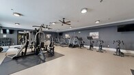 Located at 120 Chubb Ave in Lyndhurst, NJ, this fitness center provides a generously sized, single-floor workout facility designed to accommodate a full spectrum of exercise needs. The space is finished with a neutral-toned resilient floor, crisp white crown molding, and deep charcoal-gray walls that lend a modern, energizing atmosphere throughout.

The cardio zone features multiple treadmills, elliptical trainers, and stationary cycling bikes, all aligned along one wall for an organized and efficient layout. Ceiling fans with flush-mount lighting fixtures ensure consistent airflow and comfort during workouts. Wall-mounted televisions are positioned throughout the space for entertainment during cardio sessions.

The strength training area is equally well-appointed, housing a multi-station cable machine capable of a wide variety of resistance exercises. A dedicated free weights section includes a full rack of dumbbells, barbells with weight plates, kettlebells, medicine balls, and stability balls, supported by exercise benches and a vertical knee raise station. Rubber floor mats provide safety and stability in the lifting areas.

A large mirror anchors the free weights section, enhancing the sense of space and allowing for proper form checks. Natural light filters in through a window on one wall, brightening the interior. The overall layout is thoughtfully organized, making this fitness center a comprehensive and functional amenity.