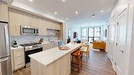 Located at 1860 Reston Row Plaza in the heart of Reston, VA, this well-appointed 3-bedroom, 2-bathroom residence offers a thoughtfully designed single-level layout with modern finishes throughout. The open-concept living room is a standout space, featuring expansive floor-to-ceiling windows that flood the room with natural light and frame views of the surrounding Reston Row neighborhood. Light wood flooring flows consistently throughout the unit, creating a cohesive and warm aesthetic. The contemporary kitchen is outfitted with light wood cabinetry, a marble-style tile backsplash, stainless steel appliances, and a central island with a white quartz countertop and seating — ideal for casual dining and entertaining. The primary bedroom is a comfortable retreat with a wall-mounted TV and direct access to a well-appointed en-suite bathroom featuring a glass-enclosed shower, a wood-toned vanity with white countertops, and an adjacent walk-in closet. A second full bathroom includes a soaking tub with marble-style tile surround and a matching light wood vanity. The additional bedrooms are bright and versatile, with access to natural light and ample space. A clean, well-lit hallway connects the rooms with easy flow. The unit's clean white walls, recessed lighting, and consistent modern design elements create a polished and functional living environment.