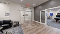 Located in Fall River, MA, this thoughtfully designed amenity space spans a generous single floor and is organized into distinct zones, each tailored for a different social experience. The centerpiece is a dedicated screening room finished in deep charcoal tones, featuring rows of plush leather recliner seats with cup holders, framed movie posters, and sleek wall-mounted sconce lighting — creating an immersive, cinema-quality atmosphere. A large-format display anchors the front wall, making it ideal for group viewings. The game room offers an expansive, open layout with patterned carpet flooring, structural columns, and recessed lighting. A full-sized pool table with pendant lighting above serves as the focal point, complemented by a wall-mounted TV, high-top seating, and a casual table-and-chair set — all framed by decorative patent art prints. The combined dining and living area transitions to warm hardwood-style flooring, providing a welcoming contrast. A substantial dining table with industrial-style metal chairs sits beneath a modern geometric pendant light, while the lounge area offers leather seating, coffee tables, and a wall-mounted TV. Glass-panel doors connect the zones, maintaining an open, airy flow throughout the space.