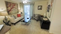 This single-floor space centers around a thoughtfully designed living room that balances comfort and style. The room features soft, neutral-toned carpeting that extends throughout, creating a warm and inviting atmosphere. A sliding glass door serves as a focal point, allowing abundant natural light to filter in through sheer, decorative curtains that frame the opening elegantly.

The living area is furnished with a cream-colored sofa accented by patterned and red throw pillows, complemented by a geometric accent chair in a neutral print. A sleek, dark coffee table with a chrome base anchors the seating arrangement, adding a contemporary touch. A matching dark console unit along one wall provides additional storage and display space.

Crown molding along the ceiling adds a refined architectural detail, while a ceiling fan with light fixture ensures both comfort and functionality. Decorative wall art, including a bold floral canvas and a framed mirror, contribute personality to the space. Overall, the living area presents a cohesive, comfortable environment suitable for both relaxing and entertaining.