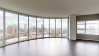 This high-rise unit is defined by its dramatic curved living area, where an expansive wall of floor-to-ceiling windows arcs gracefully to deliver breathtaking panoramic views of a vibrant city skyline. Rich dark hardwood flooring runs throughout the main living space, complementing the clean, contemporary aesthetic. The kitchen is thoughtfully appointed with crisp white cabinetry, granite countertops, a glass tile backsplash, stainless steel appliances including a range, microwave, and refrigerator, and a large central island with a built-in sink — ideal for both cooking and casual dining. A hallway with carpeted flooring connects the private areas of the unit, including bedrooms that feature large windows offering additional city views. The bathrooms are finished with light tile flooring and granite-topped vanities; one bathroom includes a soaking tub with shower combination, while another offers a walk-in shower with grab bar. A dedicated laundry area houses a stacked washer and dryer unit. Storage is well-considered, with a spacious walk-in closet providing ample rod hanging space. Throughout, neutral tones and clean lines create a calm, modern ambiance suited to urban living.