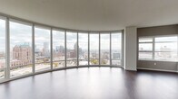 This high-rise unit is defined by its dramatic curved living area, where an expansive wall of floor-to-ceiling windows arcs gracefully to deliver breathtaking panoramic views of a vibrant city skyline. Rich dark hardwood flooring runs throughout the main living space, complementing the clean, contemporary aesthetic. The kitchen is thoughtfully appointed with crisp white cabinetry, granite countertops, a glass tile backsplash, stainless steel appliances including a range, microwave, and refrigerator, and a large central island with a built-in sink — ideal for both cooking and casual dining. A hallway with carpeted flooring connects the private areas of the unit, including bedrooms that feature large windows offering additional city views. The bathrooms are finished with light tile flooring and granite-topped vanities; one bathroom includes a soaking tub with shower combination, while another offers a walk-in shower with grab bar. A dedicated laundry area houses a stacked washer and dryer unit. Storage is well-considered, with a spacious walk-in closet providing ample rod hanging space. Throughout, neutral tones and clean lines create a calm, modern ambiance suited to urban living.