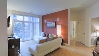 This single-floor residence offers a thoughtfully designed living space that balances modern style with everyday comfort. The living room serves as the centerpiece of the home, immediately drawing attention with a striking terracotta-hued accent wall that adds warmth and visual depth to the space. Light hardwood flooring runs throughout, complementing the neutral tones of the ceiling and trim while providing a clean, cohesive aesthetic. Floor-to-ceiling vertical blinds line an expansive window wall, allowing an abundance of natural light to flood the interior while offering adjustable privacy. The living area comfortably accommodates a generous sectional sofa arrangement, making it well-suited for both relaxing and entertaining. Built-in wall-mounted speakers suggest an integrated audio setup for an enhanced in-home experience. Artwork displayed on the accent wall adds a curated touch to the overall design. A hallway visible from the living space leads further into the residence, hinting at additional rooms beyond. The open sightlines and airy layout contribute to a sense of spaciousness throughout the home. Overall, the space strikes a balance between inviting warmth and clean, contemporary design elements.