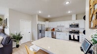 This two-story home offers a thoughtfully designed layout with functional and stylish spaces across both levels. The kitchen is a standout feature, showcasing crisp white shaker cabinetry with matte black hardware, a classic subway tile backsplash, light quartz countertops, and a full suite of stainless steel appliances including a side-by-side refrigerator with water dispenser, gas range, built-in microwave, and dishwasher. A large kitchen island provides additional prep and dining space. Light wood-look flooring flows throughout the main level, creating a bright and cohesive feel. The living room is anchored by a floor-to-ceiling stacked stone fireplace and features recessed lighting and natural light from windows with blinds and drapery. Upstairs, the primary bedroom is generously proportioned, featuring plush carpeting, large windows with plantation shutters and drape panels, and ample closet space. Additional bedrooms continue the home's clean, neutral aesthetic. A full bathroom includes a walk-in glass-enclosed shower, wood-toned vanity with granite countertop, and light hardwood flooring. The overall ambiance is warm and contemporary, with neutral tones and quality finishes throughout.