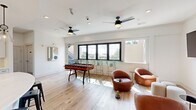 Located at 401 Oberlin in Raleigh, NC, this modern amenity space offers a well-appointed combination of interior and outdoor living areas across two levels. The main floor features an open-concept kitchen and living room finished with light hardwood-style flooring and crisp white walls throughout. The kitchen is a standout, showcasing white shaker cabinetry, quartz countertops, a herringbone-patterned tile backsplash, stainless steel appliances, and a large curved island with pendant lighting and bar seating — ideal for social gatherings. The adjoining living area is bright and contemporary, with large black-framed windows that flood the space with natural light. Dual ceiling fans, a wall-mounted flat-screen TV, comfortable leather lounge seating, and a foosball table give the room a relaxed, social atmosphere. A window-side bar counter provides additional casual seating with outdoor views. The upper level opens to a spacious rooftop terrace offering panoramic views of the Raleigh skyline and surrounding greenery. The terrace is outfitted with lounge chairs, dining sets, a turf lawn area, string lighting, and ample open-air space — making it an exceptional outdoor retreat for relaxation and entertaining.