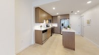 This single-floor space showcases a cohesive modern design built around a neutral palette of warm beiges, whites, and natural wood tones. Light-toned tile flooring flows consistently throughout, unifying the open layout and enhancing the overall sense of brightness.

The kitchen is a standout feature, outfitted with flat-front wood-grain cabinetry, white quartz countertops, a white subway tile backsplash, and a full suite of stainless steel appliances including a refrigerator with ice and water dispenser, electric range, over-the-range microwave, and dishwasher. A kitchen island with matching countertop provides additional workspace and casual seating potential. Recessed lighting illuminates the space effectively.

The main living area benefits from a sliding glass door that opens to an outdoor view, allowing natural light to fill the room. A ceiling fan is mounted overhead, adding both comfort and function.

The bathroom features large-format beige tile on both the floor and tub surround, a soaking tub and shower combination with a curved chrome rod and overhead rainfall-style showerhead, and recessed lighting above. The clean, minimalist design continues throughout, maintaining a consistent and polished aesthetic across the entire space.