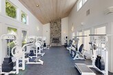 Located at 5350 N Brawley Ave in Fresno, CA, this fitness center is a thoughtfully designed single-floor facility that combines functional athleticism with striking architectural character. The space is defined by its dramatic vaulted ceiling clad in warm wood paneling, which soars overhead and creates an open, airy atmosphere that makes the room feel expansive. A floor-to-ceiling stacked stone fireplace serves as a commanding focal point, anchoring one end of the gym with a rustic yet refined aesthetic. Clerestory windows line the upper walls and large glass doors and windows run along the sides, flooding the interior with generous natural light and offering views of the surrounding outdoor greenery. The flooring transitions from durable rubber athletic matting in the main workout zone to classic white tile near the entrance, clearly delineating the activity areas. The fitness floor is well-stocked with a comprehensive array of white-framed strength training machines and cable systems, complemented by cardio equipment including treadmills, ellipticals, and stationary bikes. Wall-mounted sconce lighting adds warmth throughout, while the bright white walls enhance the sense of light and space. Double glass entry doors provide easy access and contribute to the welcoming, open feel of this well-appointed fitness facility.