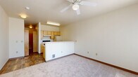 This efficient single-floor studio unit offers a well-organized open-concept layout designed to maximize functionality. The living area is carpeted and bathed in natural light from a large window fitted with vertical blinds, which provides outdoor views and is complemented by a ceiling fan overhead. The kitchen is separated from the living space by a peninsula counter, creating a natural division while maintaining an open feel. It features light wood cabinetry, an electric coil range, a refrigerator, an under-cabinet range hood, and a stainless steel sink with laminate countertops. Wood-look flooring runs throughout the kitchen, adding warmth and contrast to the carpeted living space. The hallway connects the main living area to the bathroom and includes a built-in shelving unit with wire racks, offering practical storage. The bathroom features wood-look tile flooring, clean white walls, and warm wood-toned door framing. Throughout the unit, a neutral color palette of cream and beige tones creates a clean, cohesive ambiance. The space is designed with simplicity and practicality in mind, making efficient use of every square foot.