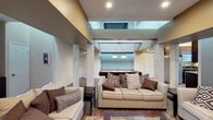 This single-floor space is thoughtfully designed to blend relaxed lounging with recreational entertainment. The living and game area serves as the central hub, anchored by a pair of plush, cream-colored sofas accented with geometric-patterned and solid brown throw pillows, arranged to encourage conversation and casual gatherings. Dark hardwood flooring runs throughout, providing a warm contrast to the neutral-toned walls and light-colored upholstery.

A standout architectural feature is the striking stepped ceiling design, which incorporates a large skylight well that draws natural light deep into the interior, creating an airy and open atmosphere despite the single-floor layout. Recessed lighting complements the natural illumination, ensuring the space is well-lit at all times.

Beyond the seating area, a full-size pool table commands attention, positioned beneath a classic pendant billiard light that adds a focal point and reinforces the entertainment-oriented character of the space. Structural columns are integrated cleanly into the design, and access to additional rooms is visible through doorways along the perimeter, suggesting a well-connected and functional floor plan throughout.