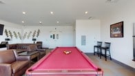 This single-floor game and lounge space is designed for recreation and socializing. The room's most prominent feature is a full-size billiard table with a bold red felt surface, positioned centrally to allow comfortable play from all sides. Recessed lighting in the white ceiling provides even, functional illumination throughout the space. To one side, a casual seating area includes dark leather lounge chairs arranged around a side table, offering a relaxed spot for guests to unwind. On the opposite side, a pair of tall bar-style chairs are positioned at a counter or high table, providing an additional seating option. The walls are painted a clean, bright white, lending an open and airy feel to the room. A decorative gold metallic wall sculpture adds an artistic focal point near the lounge area. Double white doors at the far end of the room serve as the main entry or exit, alongside a utility panel mounted on the adjacent wall. The overall aesthetic is clean and contemporary, making this a functional and inviting recreational amenity.