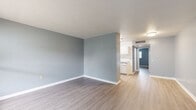 Located in Tarpon Springs, Florida, this single-story, one-bedroom, one-bathroom unit offers a well-organized layout across a compact but efficiently designed floor plan. The living room serves as the welcoming entry point, featuring warm wood-look flooring, soft blue-gray walls, and a large window that invites natural light and provides an outdoor view. The space flows into a galley-style kitchen equipped with white cabinetry, granite-style countertops, a stainless steel sink, and black appliances including a full-size refrigerator and stove — all set atop matching wood-look flooring. A hallway connects the main living areas to the private bedroom, which is a comfortable and well-proportioned room finished with the same cool blue-gray paint and wood-look flooring. The bedroom features natural light from a window and includes access to a dedicated closet with built-in shelving, offering practical storage. The bathroom is thoughtfully designed with white square wall tiles, warm mosaic tile flooring in the shower area, and a stainless steel grab bar, indicating accessibility-conscious design. Overall, the unit presents a clean, neutral aesthetic with consistent finishes throughout.