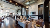 This single-floor clubhouse in Dallas, Texas, is designed with a contemporary yet refined aesthetic, blending dark espresso wood tones, stone-clad accent walls, and rich herringbone-patterned hardwood flooring throughout. The open-plan layout connects multiple functional zones seamlessly, creating a welcoming and elegant communal environment.

The central lounge area is anchored by a sleek linear fireplace set into a floor-to-ceiling limestone accent wall, topped by a wall-mounted television. Coffered ceilings with dark wood detailing add architectural depth, while geometric pendant lighting provides warm, layered illumination. Plush seating arrangements, including upholstered sofas and accent chairs, invite relaxed gathering.

Adjacent to the lounge, a well-appointed kitchen and bar area features granite countertops, dark cabinetry, open metal shelving, and pendant lighting. A large island with bar seating serves as a social hub. A separate dining area with a substantial wood table is bathed in natural light from oversized dark-framed windows that frame direct views of the outdoor pool and courtyard. The entry area further benefits from tall windows, flooding the space with sunlight and providing additional outdoor connectivity through glass sliding doors to a covered patio.