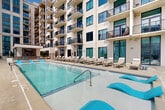 This expansive rooftop amenity deck is designed to deliver a resort-caliber experience in the heart of an urban setting. The centerpiece is a generously sized rectangular swimming pool with crystal-clear turquoise water, accented by bold blue in-pool lounge chairs that invite relaxation. Adjacent to the main pool is a separate spa, providing a dedicated space for unwinding. The pool deck is finished in large-format travertine-style tile, lending a clean and upscale aesthetic throughout the outdoor space. An abundance of cushioned chaise lounges and cabana-style seating arrangements are thoughtfully arranged along the poolside, accommodating both social gatherings and quiet leisure. A standout feature is the outdoor wet bar station, clad in rich brick with a striking hexagonal mosaic tile backsplash and topped with a mounted flat-screen television—ideal for entertaining. Wrought iron perimeter fencing with decorative stone pillars and globe lanterns frames the deck with architectural elegance while offering unobstructed panoramic views of the surrounding city skyline. The multi-story residential building rises directly behind the pool, with rows of private balconies overlooking the amenity space. This outdoor deck seamlessly blends sophisticated design with functional, resort-style living.