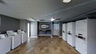This single-floor laundry facility is designed with functionality and practicality in mind. The space features a neutral color palette of gray tones on the walls, complemented by white baseboards and a textured ceiling with flush-mounted fluorescent lighting that ensures the room is well-lit throughout. The flooring consists of durable, earth-toned tile that is easy to maintain and withstands heavy foot traffic. On one side of the room, stacked washer and dryer units are neatly arranged, offering ample laundry capacity. Additional standalone washers are positioned along the opposite wall, providing further convenience for users. A built-in folding counter runs along one wall beneath a framed decorative mirror, offering a dedicated space for sorting and folding laundry. A door at the rear of the room leads to an additional area, potentially providing access to storage or utility space. Natural light filters in through a window on the side wall, contributing to the open and airy feel of the room. A fire extinguisher is visibly mounted on the wall, reflecting attention to safety standards. Overall, the space is clean, organized, and thoughtfully laid out to serve the practical needs of its users efficiently.