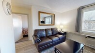 This efficiently designed one-bedroom, one-bathroom unit in Woburn, MA offers a thoughtful layout that maximizes every square foot of its single-floor footprint. The open-concept living and kitchen area serves as the heart of the unit, seamlessly blending functionality with comfort. The kitchen is well-equipped with stainless steel appliances, including a full-size refrigerator, electric range, microwave, and dishwasher, complemented by gray shaker-style cabinetry and dark granite countertops. Light hardwood-style flooring flows throughout the living area, enhancing the bright and airy atmosphere created by a large window dressed with gray curtains and blinds. The living room accommodates a leather sofa, glass coffee table, wall-mounted TV, and a small dining set, making efficient use of the shared space. A decorative gold-framed mirror adds a touch of elegance to the living area wall. The bedroom, accessed directly from the living space, features carpeted flooring, a double window with natural light, a dresser with mirror, nightstand, and a dedicated workspace with a desk. The bathroom is clean and functional, offering a white vanity with medicine cabinet mirror and a full tub/shower combination.