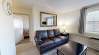 This efficiently designed one-bedroom, one-bathroom unit in Woburn, MA offers a thoughtful layout that maximizes every square foot of its single-floor footprint. The open-concept living and kitchen area serves as the heart of the unit, seamlessly blending functionality with comfort. The kitchen is well-equipped with stainless steel appliances, including a full-size refrigerator, electric range, microwave, and dishwasher, complemented by gray shaker-style cabinetry and dark granite countertops. Light hardwood-style flooring flows throughout the living area, enhancing the bright and airy atmosphere created by a large window dressed with gray curtains and blinds. The living room accommodates a leather sofa, glass coffee table, wall-mounted TV, and a small dining set, making efficient use of the shared space. A decorative gold-framed mirror adds a touch of elegance to the living area wall. The bedroom, accessed directly from the living space, features carpeted flooring, a double window with natural light, a dresser with mirror, nightstand, and a dedicated workspace with a desk. The bathroom is clean and functional, offering a white vanity with medicine cabinet mirror and a full tub/shower combination.