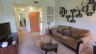 This single-floor residence offers a comfortable and thoughtfully arranged living environment with a warm, inviting ambiance throughout. The living area serves as the heart of the space, featuring plush carpeted flooring and a neutral color palette that creates a relaxed, homey atmosphere. A bold coral-toned front door makes a striking first impression, leading into an open entryway that transitions naturally into the main living space. The room accommodates a generous seating arrangement anchored by a cushioned sofa and a sleek glass-top coffee table with a contemporary sculptural base, adding a modern touch to the cozy setting. Wall décor includes a pair of framed geometric artwork pieces and an angular decorative shelf arrangement, lending character and visual interest to the space. Subtle accent lighting from flush-mount ceiling fixtures provides a soft, even glow throughout the room. The open layout between the entryway and living area promotes an easy flow and sense of openness, while the carpeted floors add warmth underfoot. Overall, the space balances practical comfort with decorative detail, offering a welcoming and functional single-level living environment.