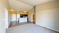 This single-floor space showcases an open-concept design that seamlessly connects the living area to the kitchen. The living space features plush carpeting in a neutral gray tone, transitioning to dark hardwood-style flooring in the kitchen and dining areas. Vaulted ceilings add a sense of openness and volume to the space, enhancing its airy feel.

The kitchen is equipped with dark cabinetry that contrasts cleanly against the white peninsula countertop, which provides both workspace and casual seating. Pendant lighting hangs above the peninsula, offering warm, focused illumination. Black appliances, including a refrigerator and microwave, complement the darker cabinetry throughout.

The walls are finished in a warm greige tone, creating a cohesive and neutral palette throughout the space. A doorway visible from the main living area leads to an additional room, suggesting a separate bedroom or hallway beyond. A second door near the kitchen area provides additional access to other parts of the unit.

Overall, the space presents a functional and modern layout with consistent finishes, practical lighting, and a clean, well-maintained aesthetic suitable for comfortable everyday living.