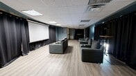 This single-floor screening and event space is designed to deliver an immersive entertainment experience within a well-appointed, intimate footprint. The room is defined by its large wall-mounted projection screen, which serves as the focal point and is flanked by floor-to-ceiling blackout curtains that line the perimeter walls. These heavy black drapes provide excellent light control and contribute to the room's theater-like atmosphere.

Seating consists of multiple sets of generously sized, upholstered sofas and lounge chairs with studded detailing and built-in cup holders, arranged in a viewing configuration facing the screen. The light wood laminate flooring provides a clean contrast to the dark curtains and teal-painted accent walls, lending the space a modern and polished aesthetic.

The drop tile ceiling houses recessed and flush-mounted lighting panels, along with a visible HVAC unit, ensuring comfortable climate control throughout. A tall bistro-style table with a pedestal base is positioned toward the rear of the room, offering a casual standing area suitable for networking or social gatherings. The overall design balances the functionality of a private screening room with the flexibility of a multipurpose event venue.