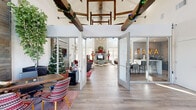 This well-appointed clubhouse offers an expansive, single-story layout designed to serve as a central hub for community living. The grand entrance opens into a soaring great room with dramatic vaulted ceilings accented by exposed rustic wood beams, wide-plank hardwood-style flooring, and a striking stone fireplace that anchors the main lounge area. A full-wall reclaimed wood feature with a mounted flat-screen TV adds warmth and character to the seating area. The adjacent Java bar is a standout feature, showcasing a bold marquee-lit "JAVA" sign set against a textured mosaic accent wall, complemented by a sleek quartz waterfall island with bar seating and built-in appliances. The fitness center is fully equipped with treadmills, cycling bikes, weight machines, and free weights, and features a glass garage door that opens to views of the outdoor pool area. A dedicated business center provides workstations, printing capabilities, and comfortable seating for productivity. A game room with a vintage pinball machine and billiards table rounds out the recreational offerings. Private office spaces with pendant lighting and carpet tile flooring offer quiet areas for focused work or meetings. Throughout, the clubhouse balances modern design with rustic, industrial touches to create an inviting and functional community space.