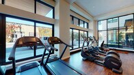 Located at 2222 NW Raleigh St in Portland, Oregon, this single-floor fitness center is a thoughtfully designed facility that blends modern aesthetics with comprehensive training capabilities. The space is bathed in natural light, courtesy of expansive floor-to-ceiling windows along multiple walls that offer views of the surrounding NW Portland neighborhood, creating an open and energizing atmosphere throughout.

The main training floor features rich hardwood flooring and soaring ceilings, lending a spacious, premium feel to the workout environment. A generous cardio zone lines the windowed perimeter, housing multiple treadmills and elliptical machines equipped with digital displays. Adjacent to this area, a strength and functional training section offers a multi-station cable machine, free weights, and specialty equipment including a RAX functional training rig. Distinctive rope pendant lighting adds an industrial-chic character to the space.

A dedicated studio area is equipped with mirrored walls, balance and gymnastics apparatus, and TRX suspension trainers, catering to a variety of training modalities. A welcoming entryway features deep navy-blue paneled wainscoting, built-in storage lockers, hanging boxing gloves, and a rack of medicine balls, setting an athletic and stylish tone upon arrival. The space is completed by a service counter area, further enhancing the facility's professional and polished character.