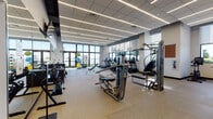 Located at 3163 SW 22nd Terrace in Miami, FL, this single-story fitness facility offers a thoughtfully designed workout environment that balances functionality with modern aesthetics. The space is characterized by a clean, contemporary architectural style, featuring a suspended tile ceiling with linear LED lighting strips that run the length of the facility, providing consistent, bright illumination throughout.

Large floor-to-ceiling windows and wide glass panels line multiple walls, flooding the interior with natural light and offering views of the surrounding outdoor greenery and Miami skyline. The flooring throughout consists of durable, speckled rubberized material suited for heavy fitness use.

The facility is organized into distinct workout zones. A dedicated strength training area houses an extensive selection of Cybex cable machines, multi-function weight stations, and free weight equipment including dumbbells and kettlebells. A separate cardio zone features a row of treadmills, ellipticals, and stair climbers positioned along the windowed walls. An open functional training area provides space for stability balls and bodyweight exercises. Full-length mirrors enhance the sense of space. Dual water fountains and direct access to an outdoor area add to the facility's convenience and overall well-appointed feel.