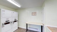 This single-story shared laundry facility is designed to serve multiple users efficiently, offering a practical and well-organized layout. The space is divided into two connected areas: a main laundry room and an entry/waiting area, both finished with light green walls and wood-look vinyl flooring that give the space a clean, understated appearance.

The main laundry area is equipped with multiple commercial-grade Speed Queen washers positioned side by side, along with stacked washer-dryer units, providing ample capacity for high-volume use. A fold-out table is available for laundry sorting and folding tasks, positioned conveniently near the machines. A baseboard heater along the wall ensures the space remains comfortable year-round. Overhead fluorescent lighting provides bright, even illumination throughout. Posted signage on the wall outlines facility rules and guidelines for users.

The adjacent entry area features a glass-panel exterior door that opens directly to an outdoor walkway surrounded by mature landscaping, allowing natural light to filter into the space. A window on the side wall further enhances the natural light. The room also includes an exit sign and wall-mounted utility controls, reflecting attention to safety and functionality throughout the facility.