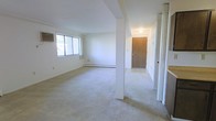 This single-floor residential space presents a clean, functional layout designed for comfortable everyday living. The open living area is the heart of the unit, characterized by wall-to-wall carpet flooring in a neutral tone that lends a warm and understated feel throughout. White walls and a simple, unadorned ceiling keep the space bright and versatile, ready for personalization. Natural light filters in through a large window on one wall, contributing to an airy atmosphere and providing a connection to the outdoors. A baseboard heating unit runs along the lower wall beneath the window, ensuring efficient climate control throughout the colder months. A structural column adds a subtle architectural element, gently defining the transition between the entryway and the main living space without fully enclosing either area. The entryway features a solid wood front door and overhead lighting, creating a practical and welcoming arrival point. A closet with bifold doors is conveniently located near the entry, offering accessible storage. The overall ambiance is modest and move-in ready, with a straightforward design that prioritizes open space and functionality.