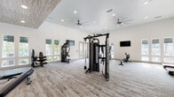 Located in Jacksonville, FL, this fitness center offers a generously proportioned, single-floor workout facility designed to support a comprehensive range of exercise routines. The space features soaring ceilings throughout, with sections highlighted by a distinctive wood-paneled ceiling accent that adds warmth and architectural character to the otherwise crisp, white interior. Large windows and glass doors flood the space with abundant natural light, providing views of the surrounding outdoor areas and creating an open, airy atmosphere. The flooring consists of durable, textured carpet throughout, lending comfort underfoot during workouts. The cardio zone is well-appointed with multiple treadmills, elliptical machines, a stair climber, and rowing machines, all positioned near windows to make the most of natural light. A dedicated strength training area features a multi-station cable machine, a Smith machine with weight plates, a full rack of dumbbells, and adjustable benches, complemented by floor-to-ceiling mirrors. A spin bike with an integrated display screen rounds out the equipment offerings. Multiple wall-mounted flat-screen TVs are positioned throughout the space, and ceiling fans ensure consistent air circulation. Built-in shelving and organized storage areas contribute to the facility's clean, functional aesthetic.