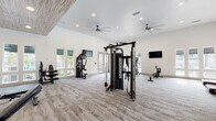 Located in Jacksonville, FL, this fitness center offers a generously proportioned, single-floor workout facility designed to support a comprehensive range of exercise routines. The space features soaring ceilings throughout, with sections highlighted by a distinctive wood-paneled ceiling accent that adds warmth and architectural character to the otherwise crisp, white interior. Large windows and glass doors flood the space with abundant natural light, providing views of the surrounding outdoor areas and creating an open, airy atmosphere. The flooring consists of durable, textured carpet throughout, lending comfort underfoot during workouts. The cardio zone is well-appointed with multiple treadmills, elliptical machines, a stair climber, and rowing machines, all positioned near windows to make the most of natural light. A dedicated strength training area features a multi-station cable machine, a Smith machine with weight plates, a full rack of dumbbells, and adjustable benches, complemented by floor-to-ceiling mirrors. A spin bike with an integrated display screen rounds out the equipment offerings. Multiple wall-mounted flat-screen TVs are positioned throughout the space, and ceiling fans ensure consistent air circulation. Built-in shelving and organized storage areas contribute to the facility's clean, functional aesthetic.
