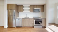 This single-floor unit combines modern design with functional living across a well-organized open layout. Warm wood-tone flooring flows consistently throughout, creating a cohesive and inviting atmosphere in every area of the space.

The living area is generously sized and bathed in natural light through two large windows that frame views of the surrounding neighborhood, including mature trees and neighboring buildings. The clean white walls and recessed lighting enhance the bright, airy feel of the room, while a wall-mounted HVAC unit ensures year-round comfort.

The kitchen is designed for efficiency and style, featuring a full suite of stainless steel appliances, including a refrigerator, electric range, over-the-range microwave, and dishwasher. Light wood-grain cabinetry lines the walls above and below the countertops, complemented by open shelving and a simple single-basin sink. The white countertops provide a clean contrast to the warm cabinetry tones.

The bathroom is compact and well-appointed, with a full bathtub and shower combination, a wall-mounted towel bar, and a vanity with wood-toned cabinetry beneath a white countertop. Recessed lighting and the continued wood-tone flooring maintain a consistent aesthetic throughout the space.