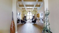 This residential community presents an impressive blend of classic architectural design and modern amenities across its single-floor clubhouse and expansive grounds. The clubhouse interior is a standout feature, showcasing a dramatic coffered ceiling with recessed lighting and an elegant chandelier that anchors the main gathering space. Arched entryways frame the room beautifully, lending a refined, traditional character throughout. Warm hardwood flooring runs the length of the space, complemented by soft sage-green walls and tasteful furnishings, including plush dark armchairs arranged around a central area rug. Decorative wall sconces, ornamental ironwork, and built-in arched niches add to the upscale ambiance. Large windows with rich drapery allow natural light to filter through, creating a welcoming atmosphere. A secondary lounge or bar area is visible to the side, offering additional seating with high-top furniture. Outdoors, the community features a generously sized swimming pool with a wide surrounding deck equipped with lounge chairs and seating areas. The pool area is enclosed by wrought-iron fencing, with mature trees and lush landscaping providing a natural backdrop. Multi-story residential buildings with balconies overlook the pool, completing the sense of a well-appointed, cohesive community setting.