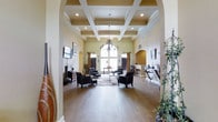 This residential community presents an impressive blend of classic architectural design and modern amenities across its single-floor clubhouse and expansive grounds. The clubhouse interior is a standout feature, showcasing a dramatic coffered ceiling with recessed lighting and an elegant chandelier that anchors the main gathering space. Arched entryways frame the room beautifully, lending a refined, traditional character throughout. Warm hardwood flooring runs the length of the space, complemented by soft sage-green walls and tasteful furnishings, including plush dark armchairs arranged around a central area rug. Decorative wall sconces, ornamental ironwork, and built-in arched niches add to the upscale ambiance. Large windows with rich drapery allow natural light to filter through, creating a welcoming atmosphere. A secondary lounge or bar area is visible to the side, offering additional seating with high-top furniture. Outdoors, the community features a generously sized swimming pool with a wide surrounding deck equipped with lounge chairs and seating areas. The pool area is enclosed by wrought-iron fencing, with mature trees and lush landscaping providing a natural backdrop. Multi-story residential buildings with balconies overlook the pool, completing the sense of a well-appointed, cohesive community setting.