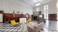 Located at 50 West 4th Street in the heart of downtown Winston-Salem, NC, this generously sized single-floor amenity space masterfully combines historic grandeur with contemporary design. The main lounge area immediately commands attention with its soaring ceilings, ornate crown molding, and richly detailed plaster walls. Rich wood wainscoting lines the walls throughout, lending warmth and a sense of institutional heritage to the space. A bold teal-blue checkered tile floor flows seamlessly across the entire floor, tying together distinct functional zones with a cohesive visual identity. The central lounge features a fireplace with an integrated media console, plush mid-century-style seating in warm gray and vibrant yellow, and elegant pendant lighting. Adjacent to the lounge, a fully equipped kitchen and bar area with teal cabinetry and counter seating provides an ideal gathering spot. A glass-partitioned area offers privacy for meetings or focused work. The space also includes a dedicated ping-pong and recreation area beneath tall windows that usher in abundant natural light, along with a fitness center accessible via a windowed door. Historical photographs and American and state flags add a meaningful nod to the building's civic past, creating an amenity floor that is as storied as it is functional.