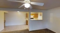 This single-floor residence offers a practical and comfortable open-concept layout designed to maximize functionality. The living area is carpeted and finished with neutral-toned walls, creating a warm and inviting atmosphere. A ceiling fan with an integrated light fixture serves as a central feature, providing both air circulation and ambient lighting throughout the space.

The kitchen is seamlessly integrated into the layout via a pass-through counter, which connects the cooking area to the living space and enhances the open feel of the home. The kitchen is equipped with warm wood-toned cabinetry, a range with an overhead ventilation hood, and a refrigerator. The cabinetry provides ample storage, while the light countertops offer generous workspace for meal preparation.

A secondary area, visible beyond a partial wall, extends the living space further, lending additional flexibility for use as a dining area or additional lounge space. The overall interior is finished in soft, neutral tones throughout, contributing to a clean and cohesive aesthetic. The layout is well-suited for everyday living, with each area flowing naturally into the next to create a connected and efficient single-floor living environment.