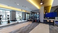 Located at 311 Karl Linn Drive in North Chesterfield, Virginia, this fitness center offers a spacious, single-floor exercise facility designed to support a comprehensive workout experience. The room is appointed with a sophisticated aesthetic, featuring a tray ceiling accented by warm amber indirect lighting that runs along the perimeter, complemented by recessed ceiling lights and ceiling fans throughout.

The floor is finished with a neutral, multi-toned patterned carpet tile that defines zones across the expansive space. Floor-to-ceiling windows line the exterior walls, flooding the interior with natural light and providing open views of an outdoor courtyard area, creating an inviting, airy atmosphere.

The cardio zone is well-stocked with elliptical trainers, stationary bikes with interactive screens, a rowing machine, and treadmills. A dedicated functional training area features a multi-station cable and resistance rig, a full rack of kettlebells and medicine balls, and TRX suspension systems. The strength training section includes a dumbbell rack with a large wall mirror, and additional cable machines for targeted muscle work.

A glass partition wall with dark metal framing divides sections of the space, lending a modern, gym-boutique feel. The overall design balances functionality with a polished, contemporary style, making this a well-rounded fitness destination.