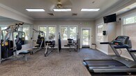 This fitness center is a thoughtfully designed, single-floor facility built to support a full range of workout routines. The space is finished with crown molding and neutral-toned walls, lending a clean and polished aesthetic throughout. Durable, textured carpet tiles cover the floors, providing both comfort and stability underfoot during exercise.

The facility is generously equipped with cardio machines, including multiple Landice treadmills and stationary bikes positioned to take advantage of the natural light streaming in through the numerous windows that line the walls. Several multi-station strength-training machines and cable systems are distributed throughout the space, accommodating a variety of resistance and weight exercises.

Large floor-to-ceiling mirrors are mounted across multiple walls, visually expanding the room and allowing users to monitor their form during workouts. The mirrors also reflect the natural light from the windows, contributing to a bright and energizing atmosphere. Overhead, flush-mounted fluorescent light panels and skylight-style ceiling fixtures ensure consistent, even illumination throughout the day.

Interior glass-panel doors connect sections of the facility, maintaining an open flow between areas. The space benefits from multiple exterior-facing windows and glass doors that offer views of surrounding greenery, creating an inviting environment. Wall-mounted televisions provide additional entertainment options for users during their sessions.