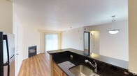 This single-floor residence offers an open and functional layout that seamlessly connects the kitchen and living areas. The kitchen is anchored by rich, dark granite countertops that extend across an L-shaped peninsula, providing generous prep space and a natural divider between the cooking and living zones. Warm honey-toned cabinetry below complements the countertops, and a stainless steel sink with a classic faucet adds a practical finishing touch.

The living area features warm hardwood flooring that runs throughout, contributing to a cohesive and inviting atmosphere. A fireplace with a sleek, dark surround serves as a focal point along one wall, adding both warmth and character to the space. Vertical blinds cover a large sliding glass door or window, allowing natural light to fill the room while offering privacy control.

Neutral, light-toned walls and a textured white ceiling keep the space feeling bright and airy. A doorway visible toward the rear of the unit leads to additional rooms, with wall-mounted lighting adding a subtle accent. Overall, the residence balances practical design with comfortable, everyday livability.