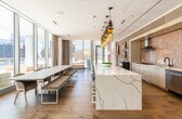 This single-floor space is a modern, upscale gathering venue defined by its striking architectural design and versatile layout. Floor-to-ceiling glass walls and sliding glass partitions run along the perimeter, flooding the interior with natural light and offering panoramic views of the surrounding cityscape. Light-toned hardwood flooring flows consistently throughout, tying together the various functional zones.

The main kitchen area is a standout feature, equipped with professional-grade stainless steel appliances, wall ovens, a gas cooktop, and a large waterfall-edge marble island beneath a dramatic cluster of multicolored pendant lights. A decorative mosaic tile backsplash adds visual texture behind the cabinetry. Adjacent to the main kitchen is a secondary wet bar area complete with a draft beer tap system, gas range, range hood, and a textured stone tile surround — ideal for catered events.

The expansive lounge area is anchored by a floor-to-ceiling travertine stone fireplace with a linear gas insert. Flexible, colorful modular seating and a sculptural cylindrical pendant light give the space an artistic, inviting character. A billiards table and sheer floor-length curtains add to the relaxed yet refined ambiance.

Direct access to an outdoor terrace is available through multiple glass doors, seamlessly extending the gathering space outdoors with open city views.