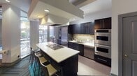 Located in Charlotte's South End neighborhood, this single-floor amenity space at 222 E Bland St offers a thoughtfully designed environment that blends social, culinary, and collaborative functions. The entrance welcomes visitors with a curated lounge area furnished with plush sofas and armchairs accented by teal and yellow throw pillows, anchored by a bold portrait artwork and recessed lighting overhead. Pendant globe lights illuminate a secondary seating area near glass double doors that open directly to an outdoor patio, flooding the space with natural light and views of the surrounding streetscape. The fully equipped kitchen is a standout feature, showcasing dark espresso cabinetry, a mosaic tile backsplash, sleek quartz countertops, a gas cooktop island with bar seating, stainless steel appliances including a double wall oven and built-in refrigerator, and a dedicated sink station. Adjacent to the lounge, a co-working and dining zone features marble-top high tables paired with contemporary bar-height chairs, wall-mounted flat-screen televisions, and frosted window partitions for a semi-private atmosphere. Throughout the space, vibrant geometric patterned carpet tiles in teal, green, and grey tie the distinct zones together, creating a cohesive and energetic aesthetic suited for both gathering and productivity.