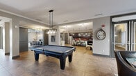 This well-appointed amenity space spans a single floor and is thoughtfully designed to serve both recreational and functional needs. The open layout flows seamlessly between distinct zones, each with its own character and purpose.

At the heart of the space is a game room anchored by a full-size pool table beneath a modern pendant chandelier, set on geometric patterned hardwood-style tile flooring. Adjacent to it, a fully equipped kitchen features floor-to-ceiling dark espresso cabinetry, a striking red tile backsplash, granite countertops, stainless steel appliances, and a large island with seating — perfect for casual gatherings or catered events.

A formal dining area with a tray ceiling, geometric-patterned carpet, and a statement chandelier provides an elegant setting for group meals or meetings. Natural light filters in through large windows with roller shades, creating a bright and welcoming atmosphere.

The business center offers a granite-topped workstation with computers and printing equipment, accented by decorative pendant lighting and bold patterned wallpaper. A private lounge or card room, accessible through glass-paneled gray doors, features a textured metallic tile accent wall with a mounted TV, offering an intimate retreat.

A cozy seating nook near French doors opens to an outdoor patio, connecting the interior to the outdoors with ease. Throughout, consistent use of dark trim, industrial-style lighting fixtures, and curated décor creates a cohesive, upscale ambiance.