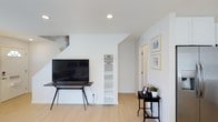 This thoughtfully designed two-floor space embraces a minimalist aesthetic, characterized by crisp white walls, light wood-tone flooring, and a clean, uncluttered layout that promotes a sense of openness and calm. The main living area is bright and welcoming, featuring recessed ceiling lighting that evenly illuminates the room without disrupting the sleek overhead finish. A staircase leads to the upper floor, adding vertical dimension to the space and serving as a subtle architectural focal point. The living area comfortably accommodates a media setup, with ample wall space ideal for display and décor. A built-in vertical HVAC unit is neatly integrated into the wall, maintaining the room's streamlined appearance. A hallway leads further into the space, offering access to additional rooms, with framed wall art adding a personal and curated touch to the corridor. The overall design philosophy prioritizes simplicity and functionality, with neutral tones and natural-feeling materials creating a cohesive, harmonious environment throughout. The combination of recessed lighting, light flooring, and white walls ensures the space feels bright and airy at all times, making it both practical and visually appealing.