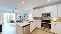 Located at 5948 Aretha Place in Virginia Beach, VA, this single-floor, 1-bedroom, 1-bathroom residence offers a thoughtfully designed open-concept layout with modern finishes throughout. The kitchen is a standout feature, showcasing crisp white shaker cabinetry, a neutral tile backsplash, a peninsula with a built-in sink and breakfast bar seating, and a full suite of stainless steel appliances including a refrigerator, range, microwave, and dishwasher. The kitchen flows seamlessly into the living room, which is brightened by recessed lighting, a ceiling fan, and a sliding glass door leading to a private balcony with views of the surrounding community. The bedroom is a comfortable retreat finished with plush carpeting, neutral-toned walls, and a window that allows natural light to fill the space. The full bathroom features wood-look luxury vinyl flooring, white subway tile surrounding a soaking tub and shower combination, a white vanity with quartz countertop, and gold-toned hardware accents. A dedicated laundry area houses a stacked washer and dryer unit with additional shelving for storage. The home's clean, contemporary aesthetic is consistent throughout, making it a functional and inviting living space.