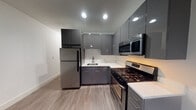 Located at Village Place on West Main Street in Bay Shore, New York, this single-floor unit offers a thoughtfully designed layout with modern finishes throughout. The kitchen is a standout feature, equipped with sleek, high-gloss gray cabinetry extending from floor to ceiling, white quartz countertops, stainless steel appliances including a gas range, over-the-range microwave, and refrigerator, as well as recessed lighting that illuminates the space efficiently. The light wood-tone flooring in the kitchen transitions seamlessly into the open living area, which features durable, dark wood-tone hardwood-style flooring. The living space is bright and airy, benefiting from a large window that allows natural light to filter in, complemented by a ceiling fan with an integrated light fixture. A wall-mounted HVAC unit ensures year-round climate comfort. The hallway connecting the rooms is clean and minimal, with crisp white walls, solid panel doors, and consistent flooring that ties the layout together. The bathroom is finished with bright white tile and features a walk-in glass-enclosed shower with polished chrome hardware and handheld fixtures, conveying a spa-like cleanliness. Overall, the unit presents a contemporary, move-in-ready aesthetic with cohesive design elements throughout.