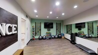 This single-floor fitness space is thoughtfully designed to support a comprehensive workout experience. The room features a clean, modern aesthetic with white ceilings lined with recessed lighting that ensures bright, even illumination throughout. A signature sage-green accent wall serves as the focal point, anchoring a wall-mounted flat-screen TV that allows for guided workout sessions or entertainment during exercise.

The space is outfitted with a variety of functional training equipment, including two sets of wall-mounted wooden stall bars ideal for stretching, mobility work, and bodyweight exercises. A rack of medicine balls in assorted weights lines one wall, complemented by a set of stability and exercise balls arranged along the base of the accent wall. A tiered dumbbell rack positioned near the mirrored wall provides a wide range of free weight options for strength training.

The full-length mirror along one wall enhances the sense of space and allows users to monitor their form during workouts. Durable wood-look flooring adds warmth while offering a practical, easy-to-clean surface suited for physical activity. An adjacent accessible restroom adds convenience to the facility. Overall, this fitness room combines functionality with a welcoming atmosphere, making it suitable for a diverse range of exercise routines.